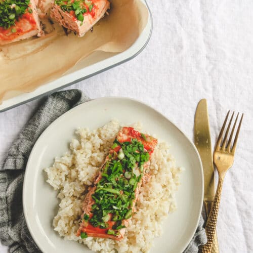 mayo ketchup baked salmon with fresh herbs on top serve over rice in a white plate with fork and knife on the side.