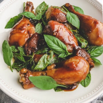 ooked three cup chicken drumsticks with basil in a white plate on top of gray cloth.