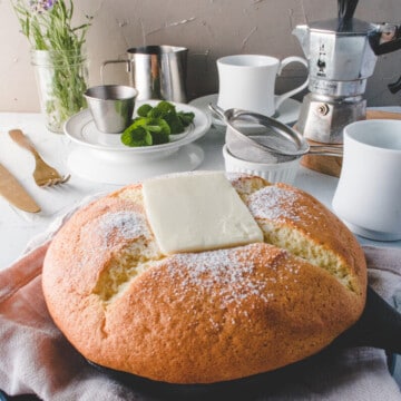 castella pancake in a black skillet top with a piece of butter. cups, plates, coffee maker, fork, knife, sifter and plates on the side.