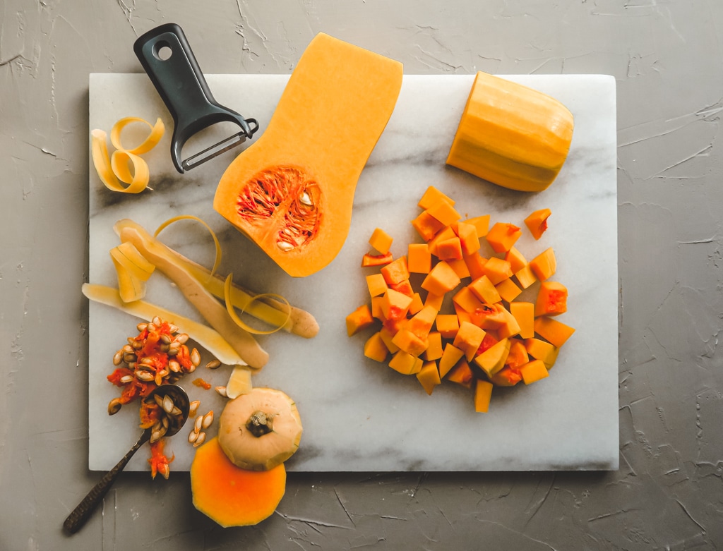 white marble cutting board with butternut squash cut up in cubes and skinned in halves.