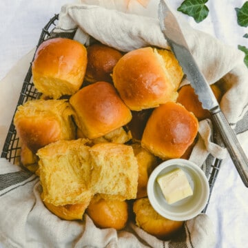 yellow bread rolls, butter and butter knife in a black basket lined with dish towel.