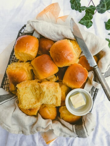 yellow bread rolls, butter and butter knife in a black basket lined with dish towel.