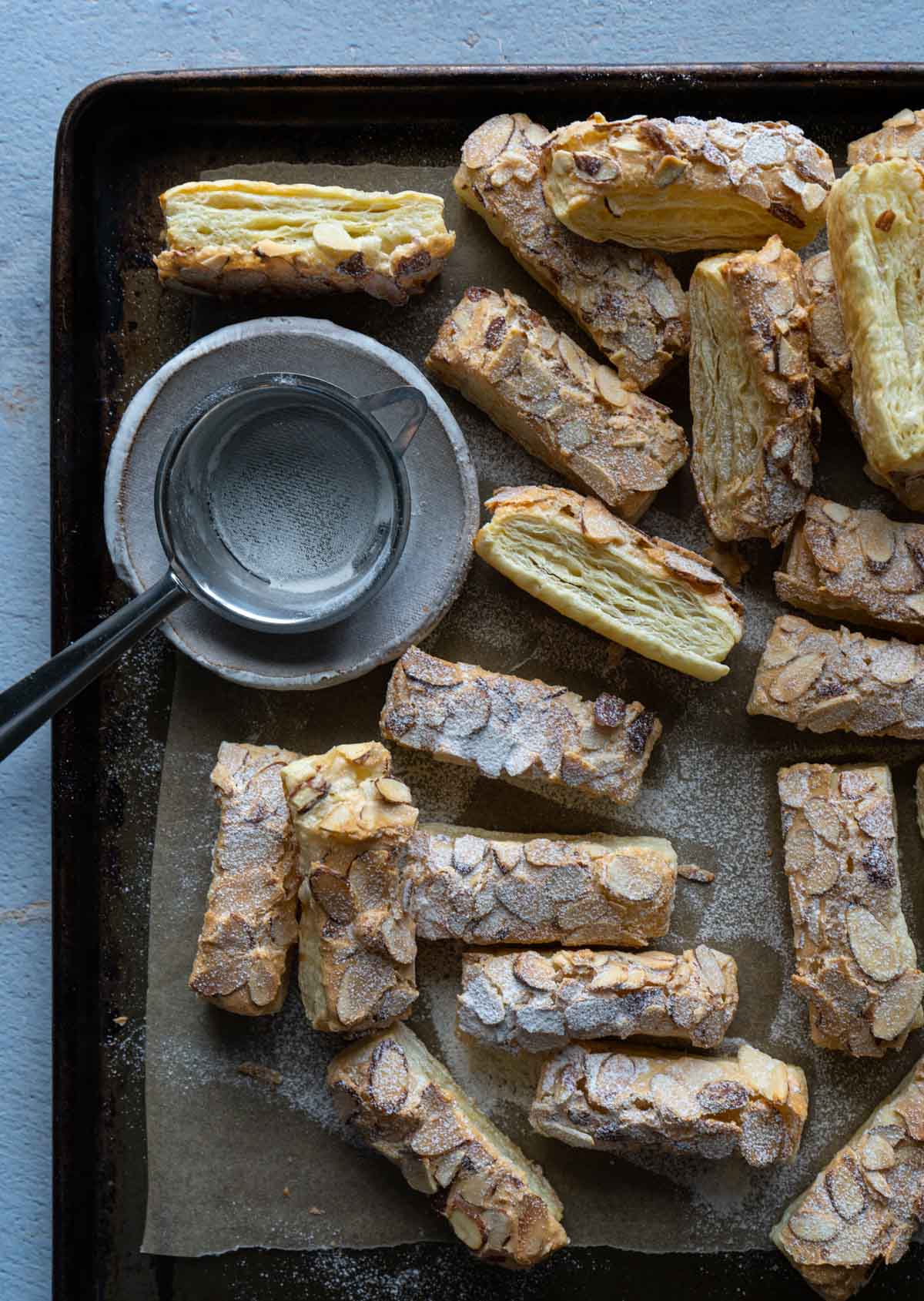 almond puff pastry cookies in a baking tray with a small plate and sifter on the side.