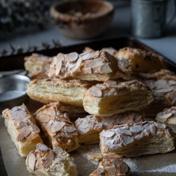 almond puff pastry cookies in a baking tray with a wood bowl. plant and a grey cup.