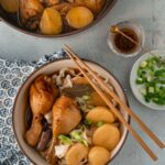 A pot and a bowl of chicken stew with potatoes and noodles, a small dish with chopped scallion and a pinch bowl with chili oil on the side.