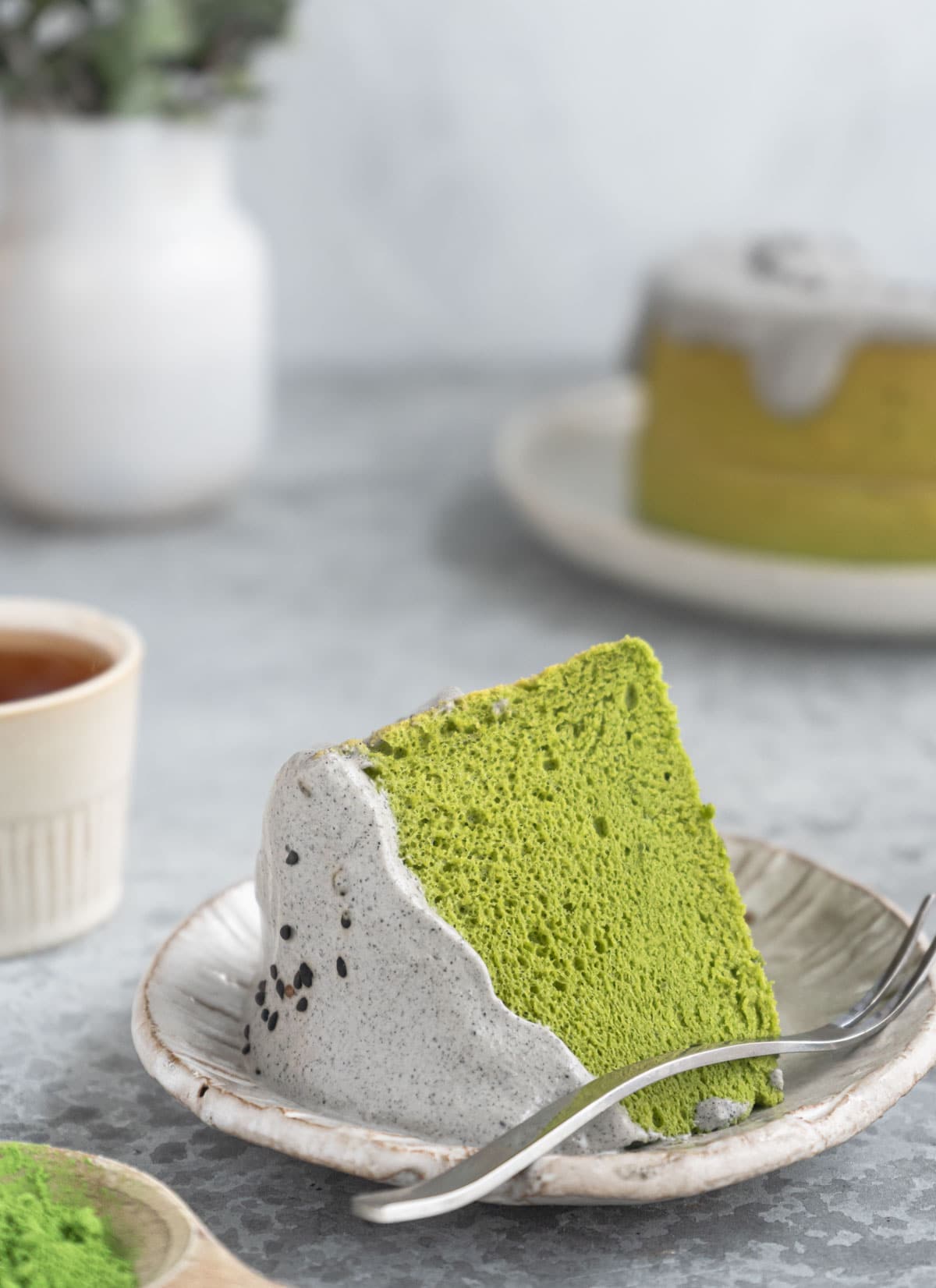 A piece of matcha chiffon cake with black sesame cream,, a cup of tea and a vase.