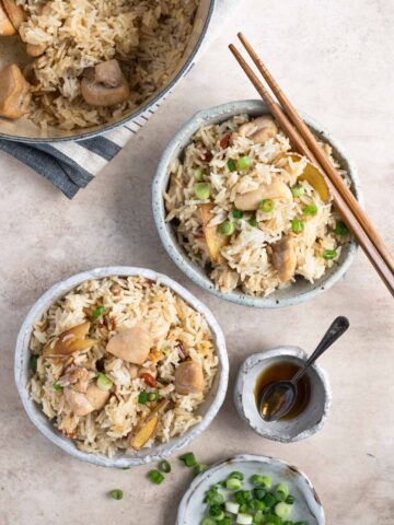 Two bowls of Taiwanese sesame oil chicken rice along with Dutch oven, a pinch bowl of sesame oil and a small plate of chopped scallions on the side.