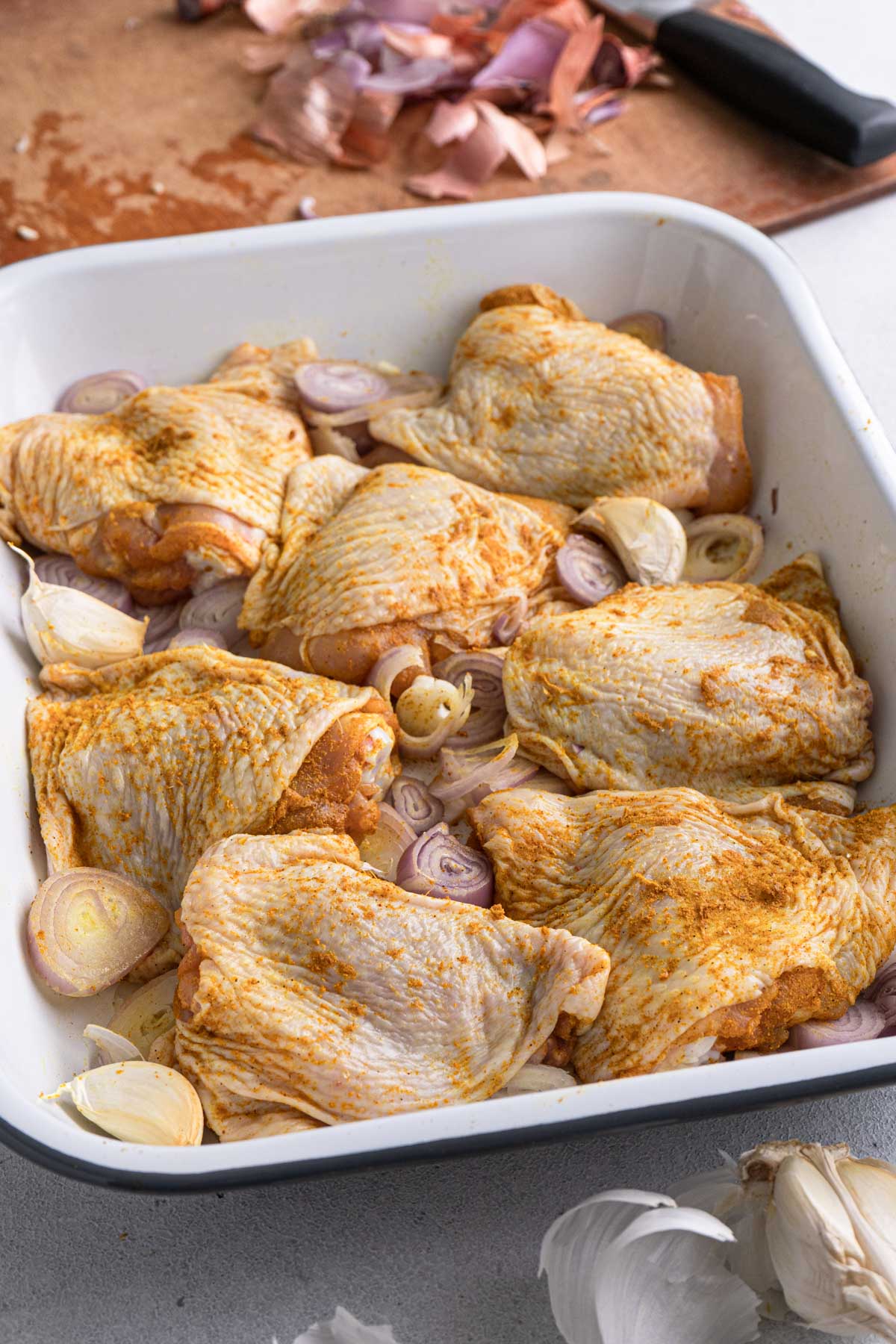 A tray of uncooked chicken thigh with shallot slices and garlic cloves. 