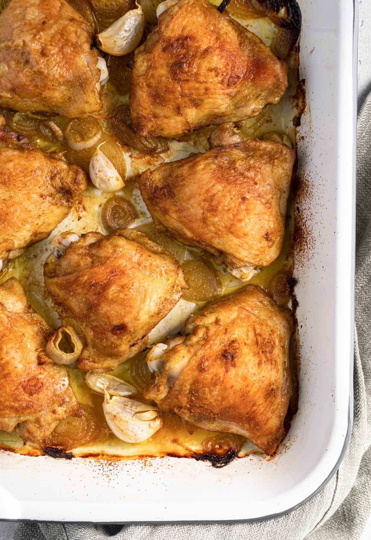 Baked chicken thighs with garlic and sliced shallot on a white tray.