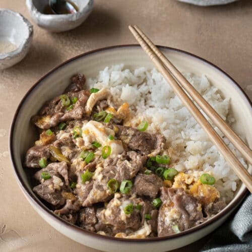 A bowl of beef and egg over rice with a pair of chopsticks on it.