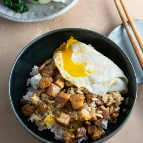 A bowl of Taiwanese braised pork rice with an over easy egg, a pair of chopsticks and a plate of vegetable on the side.