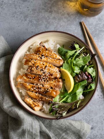 A bowl of teriyaki chicken over rice with green salad, a pair of chopsticks and a cup of tea.