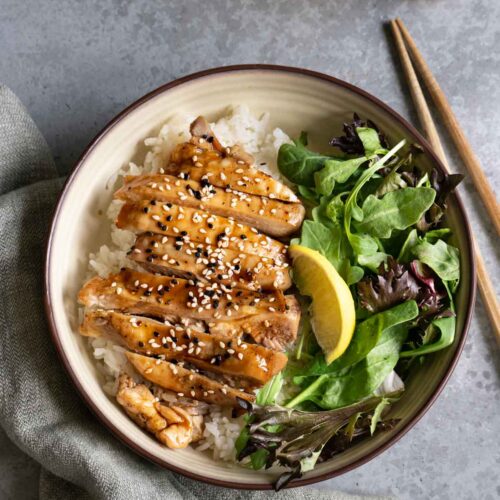 A bowl of teriyaki chicken over rice with green salad, a pair of chopsticks and a cup of tea.