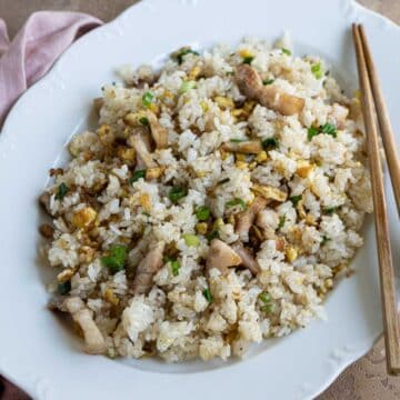 Pork belly fried rice with egg and scallion in a white oval plate with a pair of chopsticks, pink napkins and a small plate of chopped scallion on the side.
