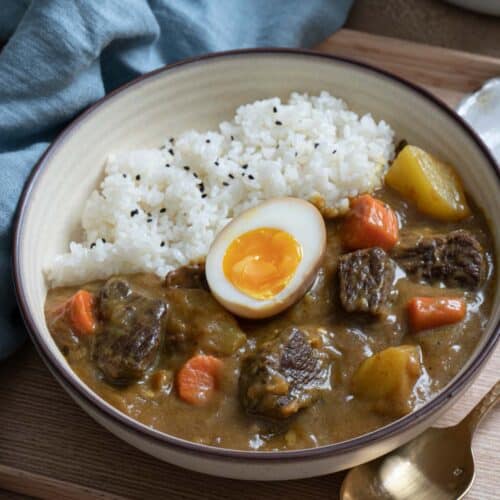 A bowl of Japanese beef curry with rice and half of a soft boil egg, with a blue towel on the side.