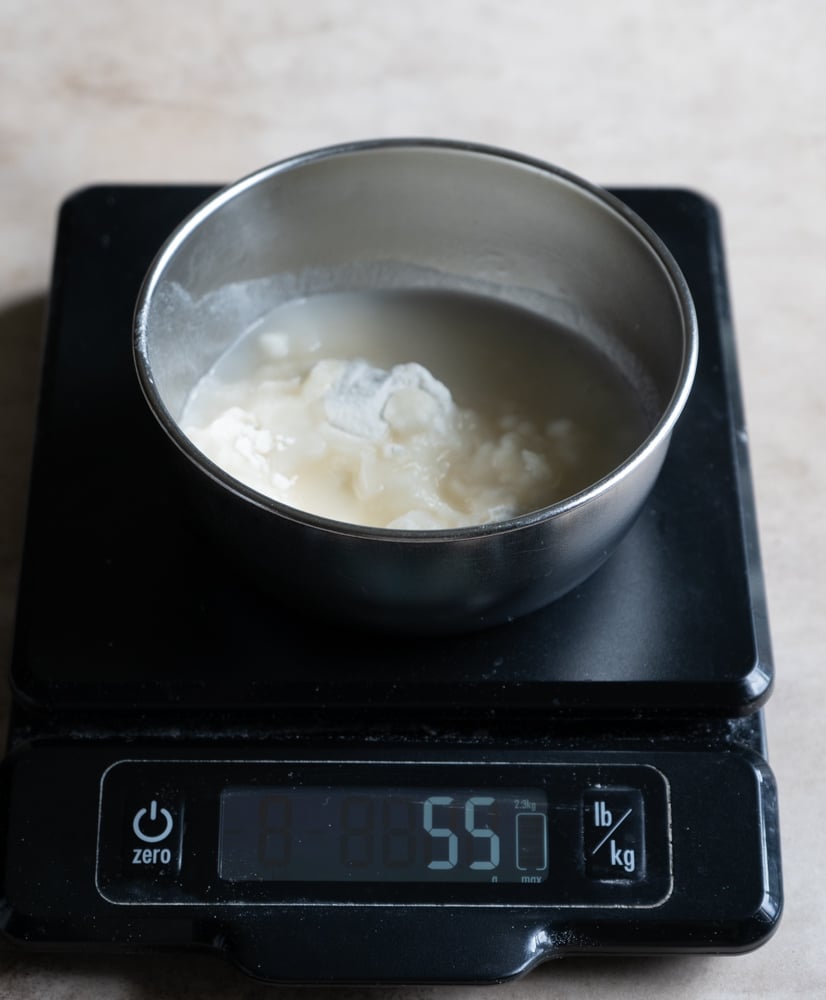 A metal bowl containing a thick white flour-water mixture (tangzhong) sits on a digital kitchen scale, which displays a weight of 55 grams.