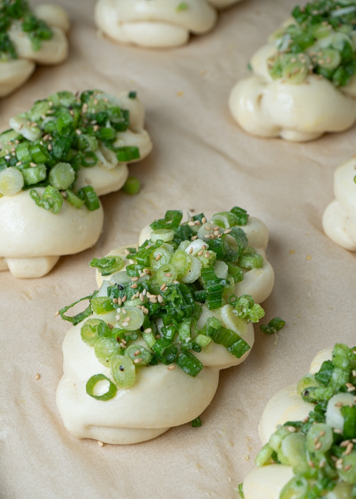 Rows of braided, unbaked dough topped with chopped scallions and sesame seeds on a parchment-lined baking sheet.