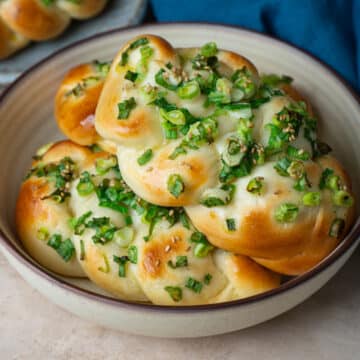 A bowl filled with freshly baked Taiwanese scallion bread, braided and golden brown, topped with chopped green onions and sesame seeds, set on a neutral-toned surface with a blue cloth napkin nearby.