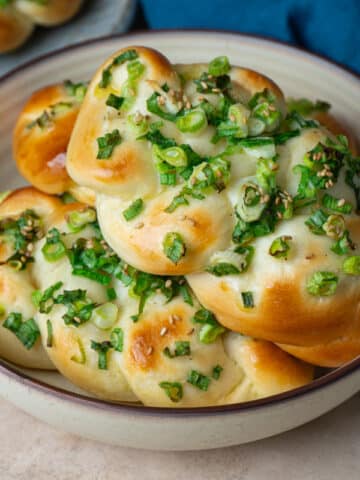 A bowl filled with freshly baked Taiwanese scallion bread, braided and golden brown, topped with chopped green onions and sesame seeds, set on a neutral-toned surface with a blue cloth napkin nearby.