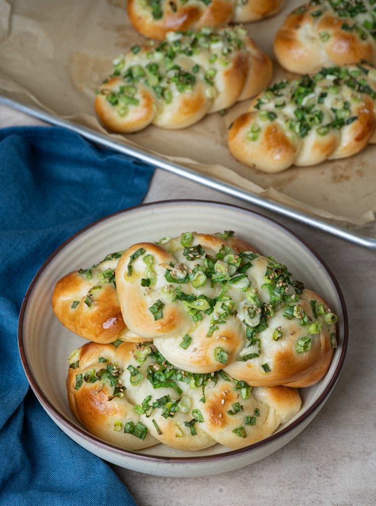 Three golden-brown scallion buns in a ceramic bowl with a blue napkin on the side and more breads on the baking tray lined with parchment paper. 