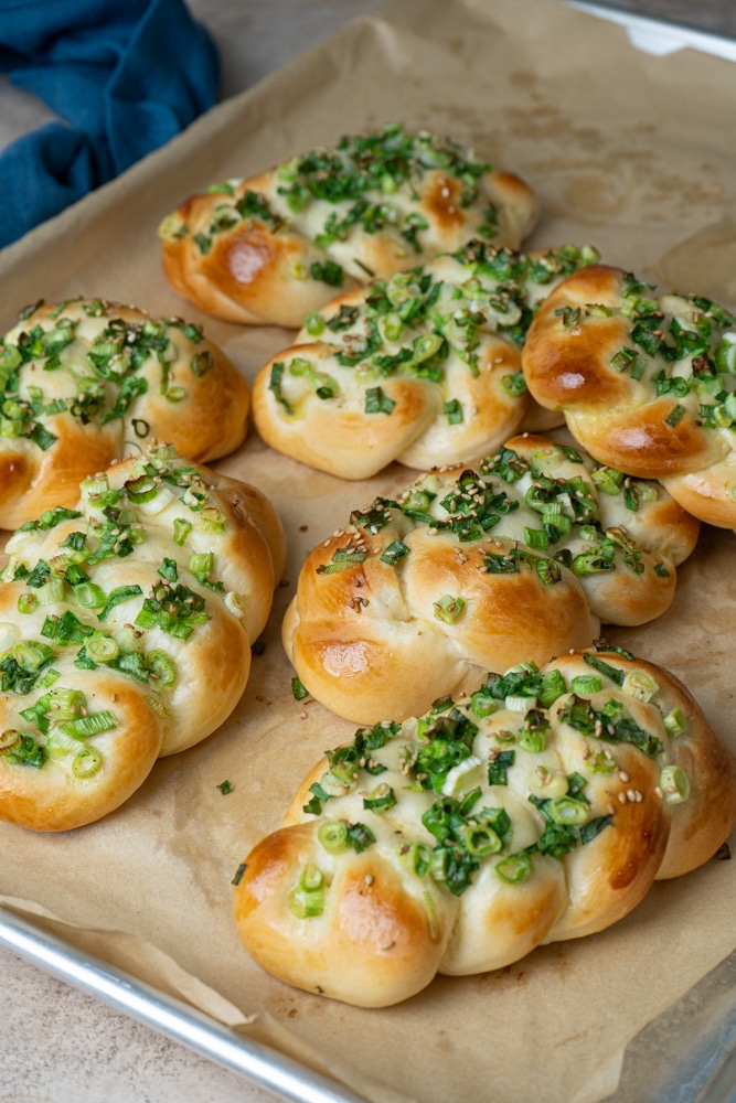 Freshly baked Taiwanese scallion breads arranged on a parchment-lined baking sheet. The golden-brown braided buns are topped with chopped green onions and sesame seeds, with a blue cloth partially visible in the background.