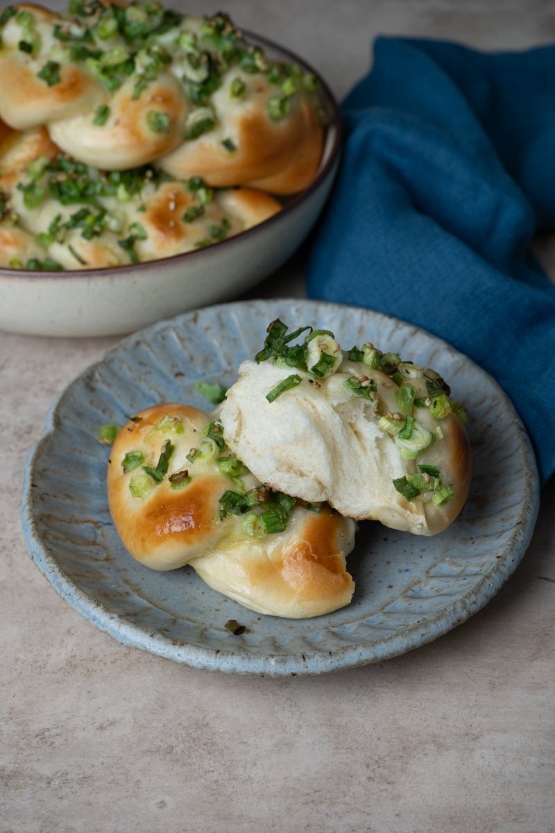 A close-up of two golden-brown scallion buns on a ceramic plate, one of them torn open to show the soft, fluffy interior, with more buns in a bowl in the background.