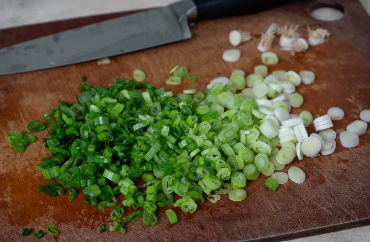 A cutting board with freshly chopped scallions in a mix of green and white segments, with a large knife placed above them.