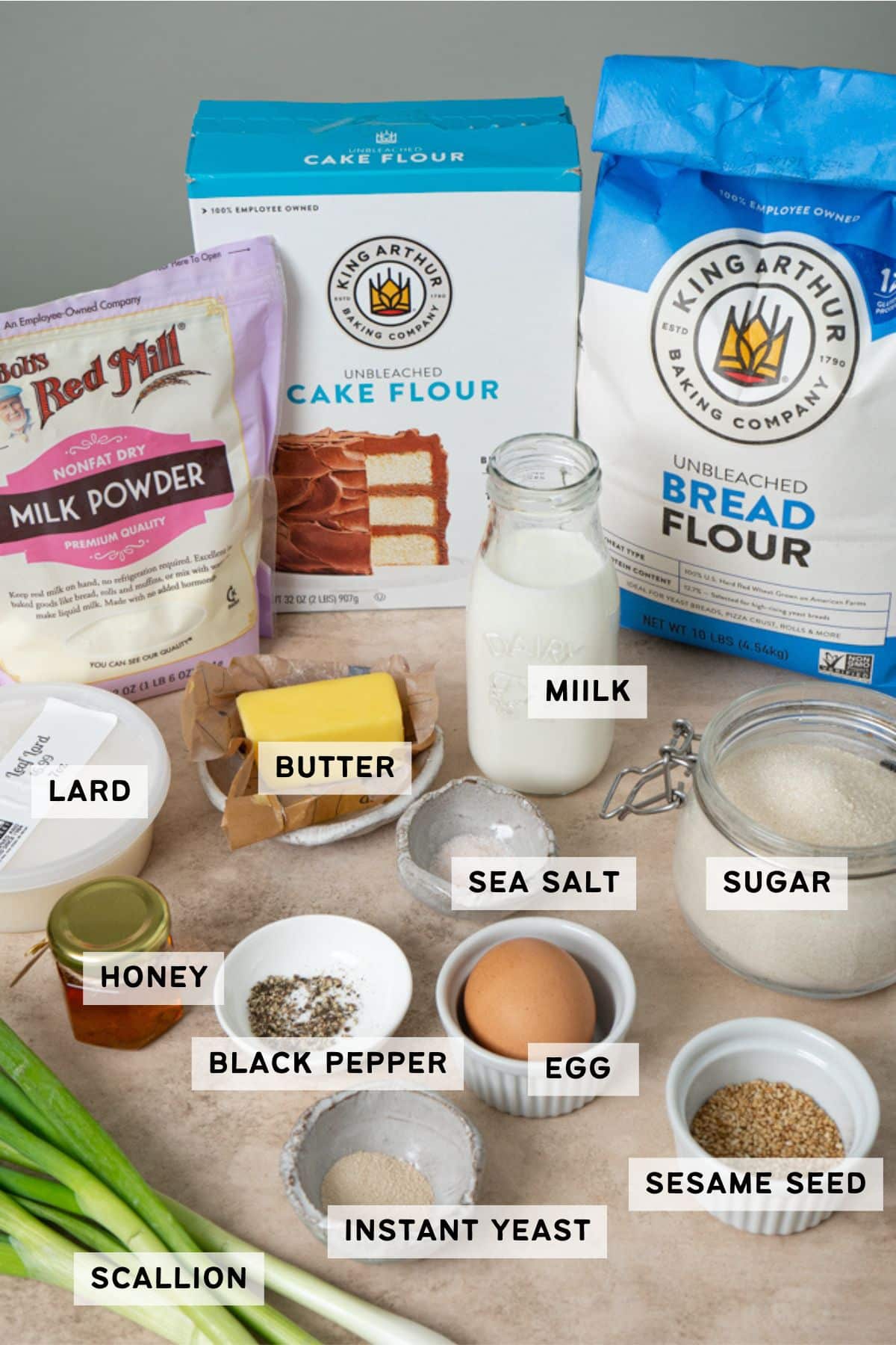 Flat lay of ingredients for scallion bread, including bread flour, cake flour, milk powder, Instant yeast, sugar, salt, milk in a small cup, scallions, lard, egg and sesame seeds. 