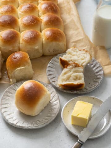 Tray of freshly baked dinner rolls with shiny golden crusts, one roll torn open on a plate to show the light, fluffy interior, served with butter and milk.