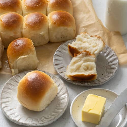 Tray of freshly baked dinner rolls with shiny golden crusts, one roll torn open on a plate to show the light, fluffy interior, served with butter and milk.