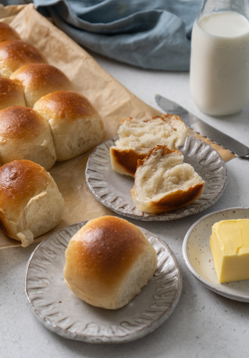 Tray of freshly baked dinner rolls with shiny golden crusts, one roll torn open on a plate to show the light, fluffy interior, served with butter and milk.