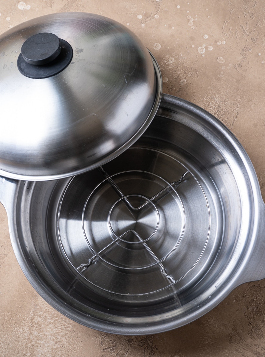 A stainless steel steamer pot with a wire rack inside and a partially open lid, placed on a beige countertop.