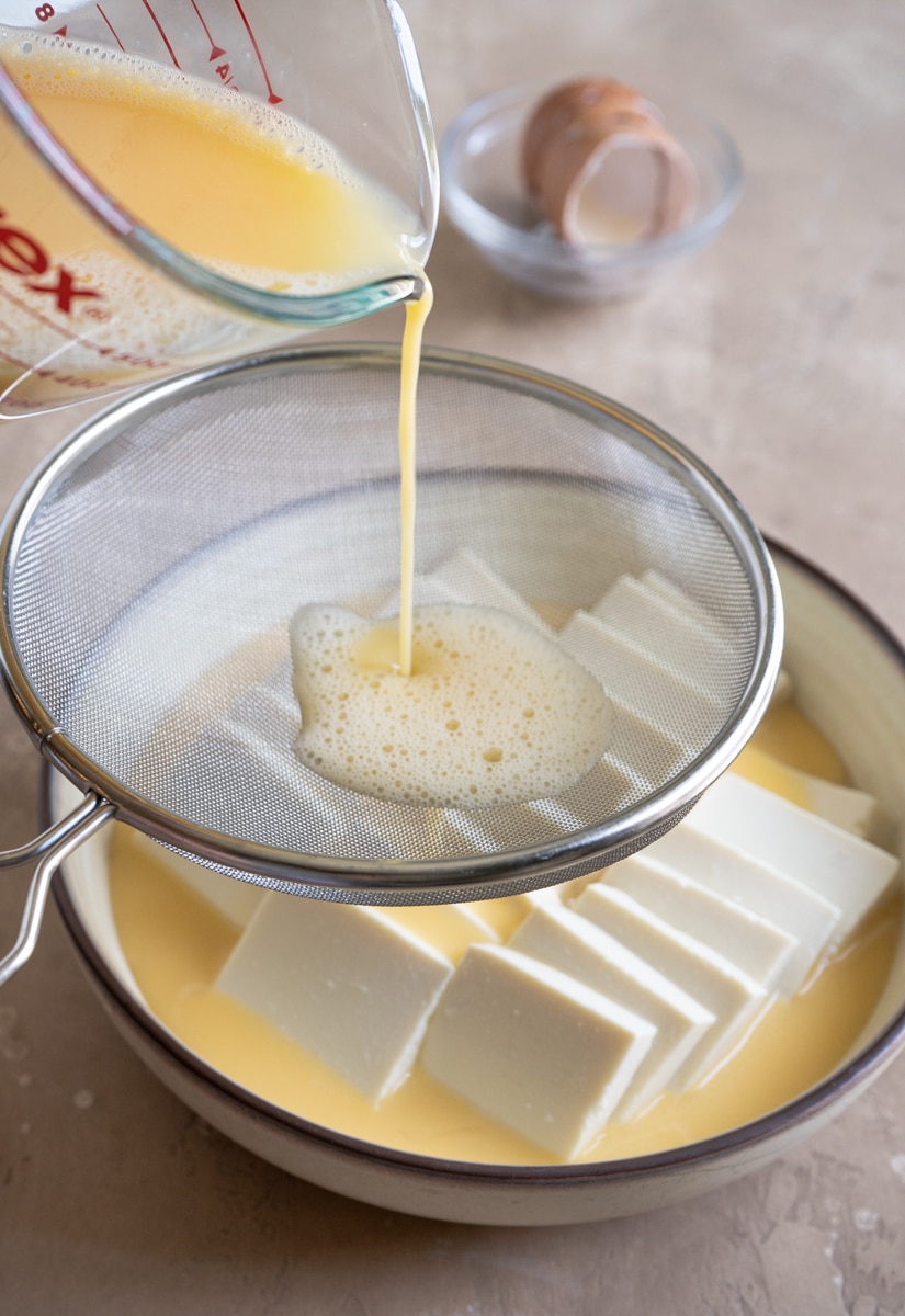 Egg mixture being poured through a fine mesh strainer over sliced tofu in a bowl, preparing for steaming.