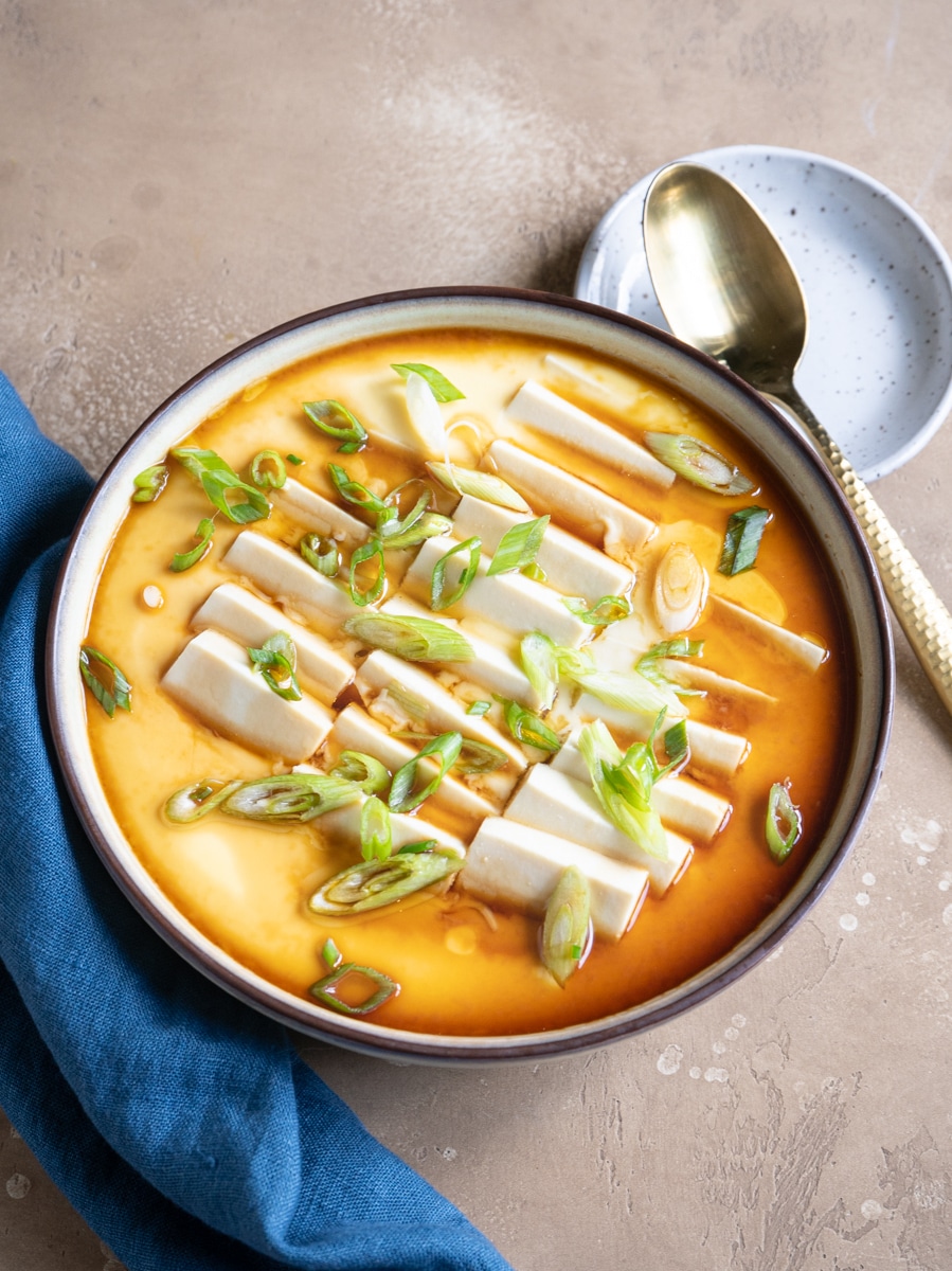 A bowl of steamed egg custard topped with rectangular slices of tofu and garnished with sliced green onions. The dish is presented on a beige surface with a blue napkin, a gold spoon, and a small plate beside the bowl.