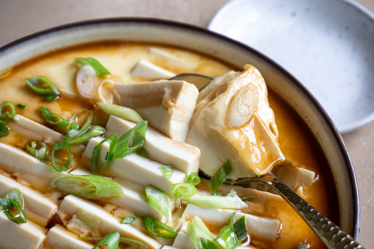 Close-up of steamed egg and tofu dish topped with sliced green onions and drizzled with soy sauce, with a spoon lifting a tender piece.