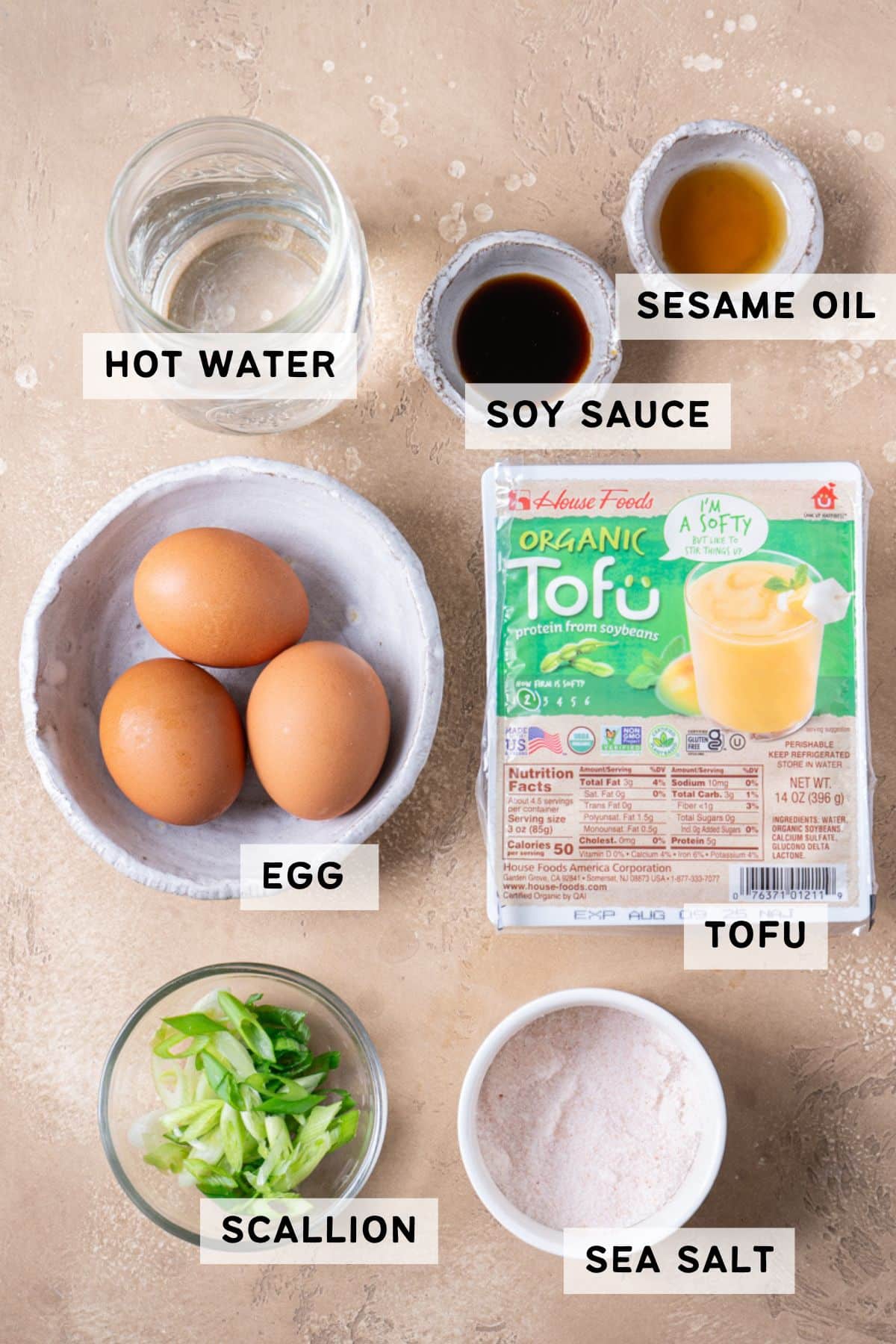 An overhead view of labeled ingredients for steamed egg with tofu, including a jar of warm water, a small bowl of soy sauce, a small bowl of sesame oil, three eggs in a bowl, a package of organic tofu, a bowl of sliced scallions, and a bowl of sea salt, all arranged on a beige surface.