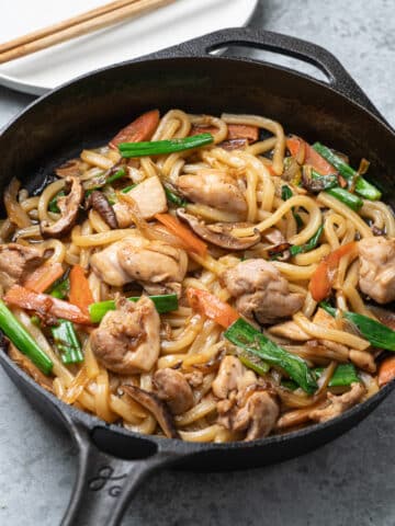 A cast iron skillet filled with stir-fried chicken udon noodles, featuring pieces of chicken, sliced carrots, green onions, and mushrooms in a glossy soy-based sauce.