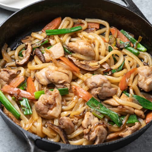 A cast iron skillet filled with stir-fried chicken udon noodles, featuring pieces of chicken, sliced carrots, green onions, and mushrooms in a glossy soy-based sauce.