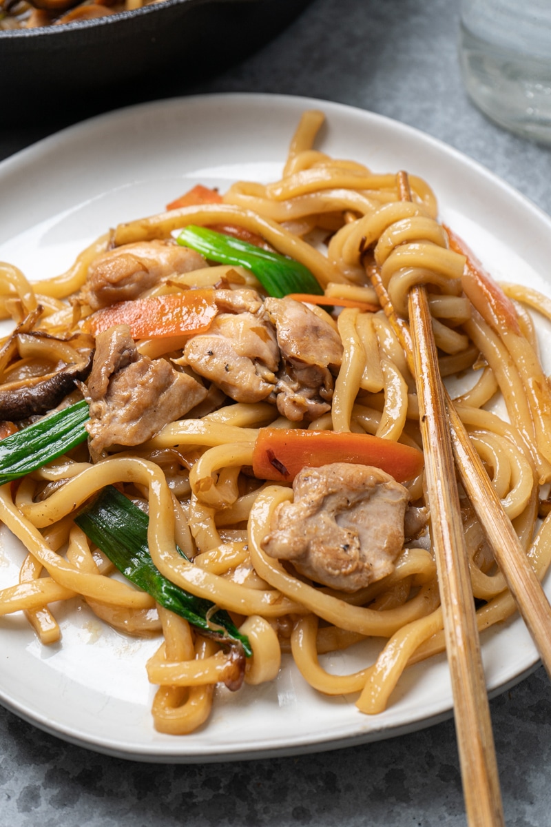 Close-up of a plate of stir-fried chicken udon noodles with pieces of chicken, carrots, green onions, and mushrooms, with chopsticks picking up a portion of the glossy noodles.