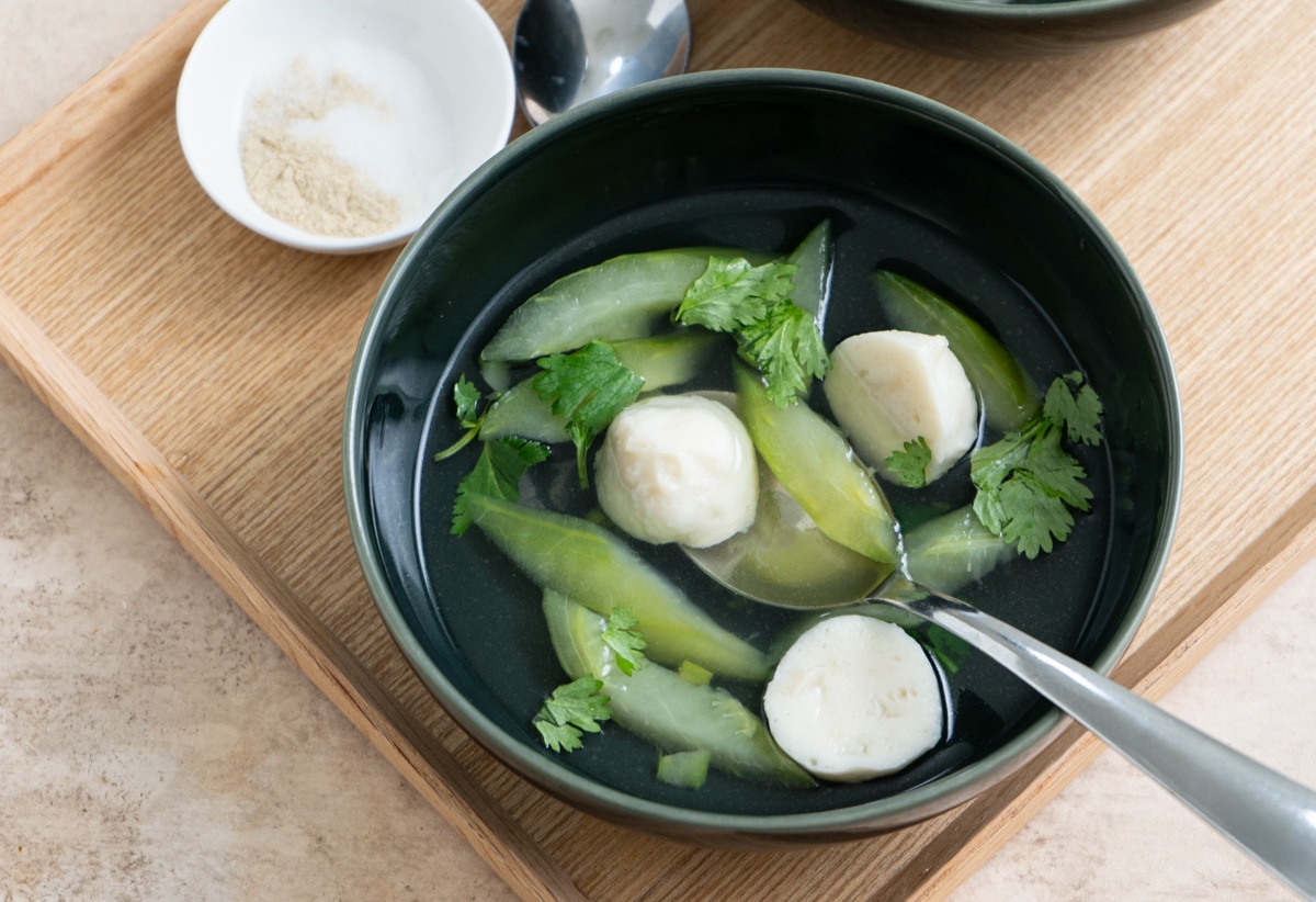 A bowl of Chinese cucumber soup with fish balls and cilantro on a wooden tray. with a spoon inside the bowl, and a small plate of salt and white pepper powder on the side.