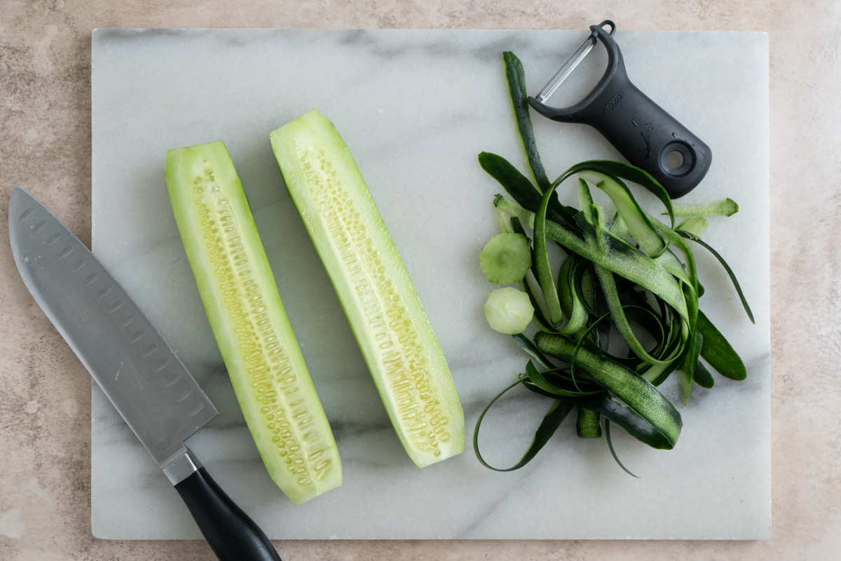 Peeled cucumbers sliced lengthwise on a marble cutting board, with a knife, vegetable peeler, and a pile of cucumber peels beside them.