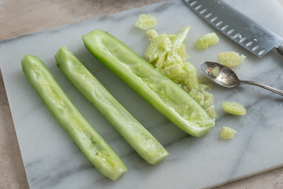 Halved cucumbers with their seeds being scooped out on a cutting board, next to a spoon, seeds, and a knife.