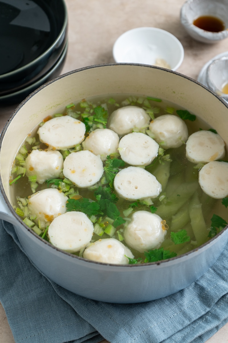 A pot of Chinese cucumber and fishball soup simmering, with sliced cucumber, fishballs, chopped cilantro, and seasonings floating in a clear broth.