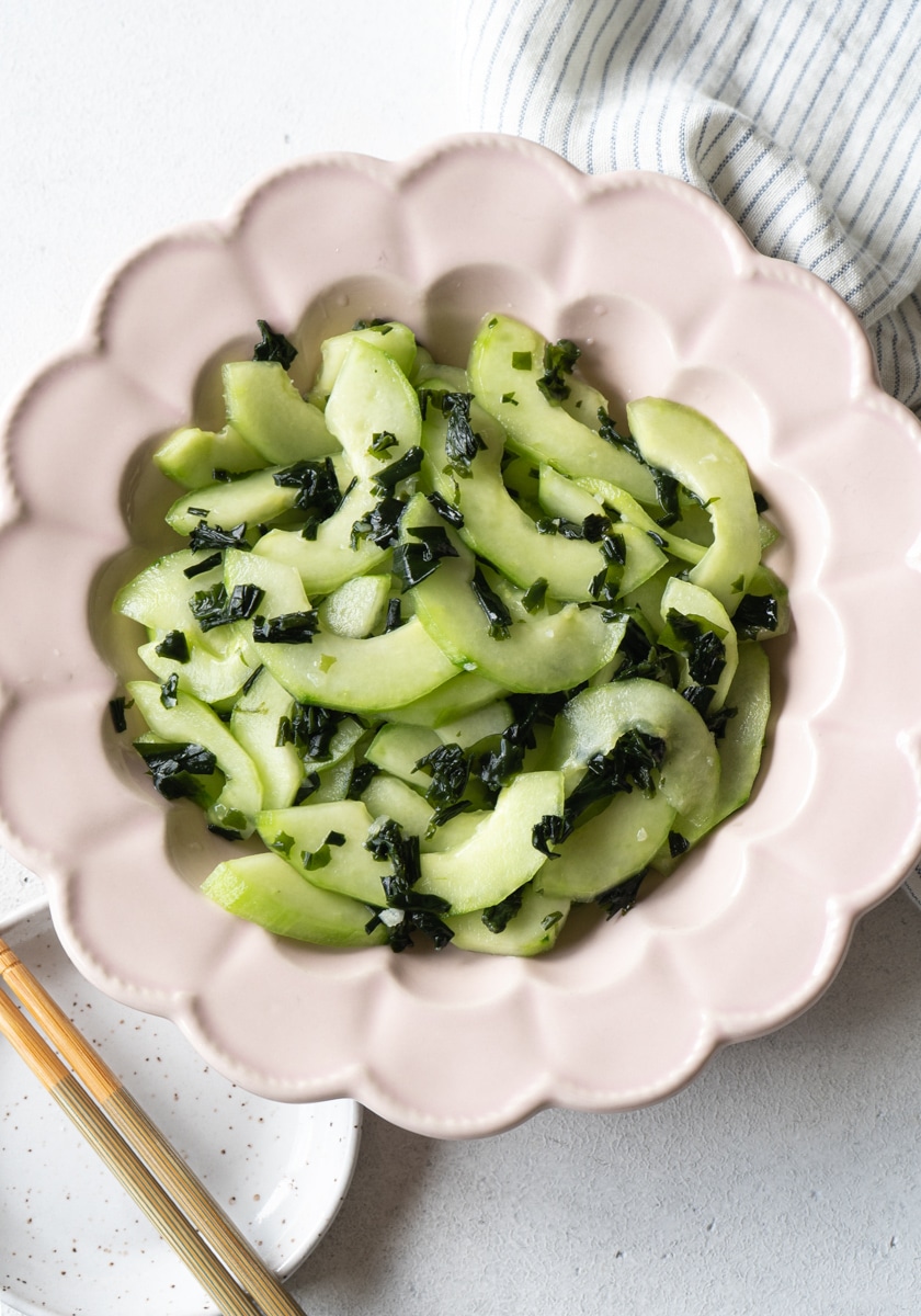 sliced cucumber with seaweed pieces in a pink flower shape plate, and a pair of chopsticks on the lower left corner on top of a small white plate.