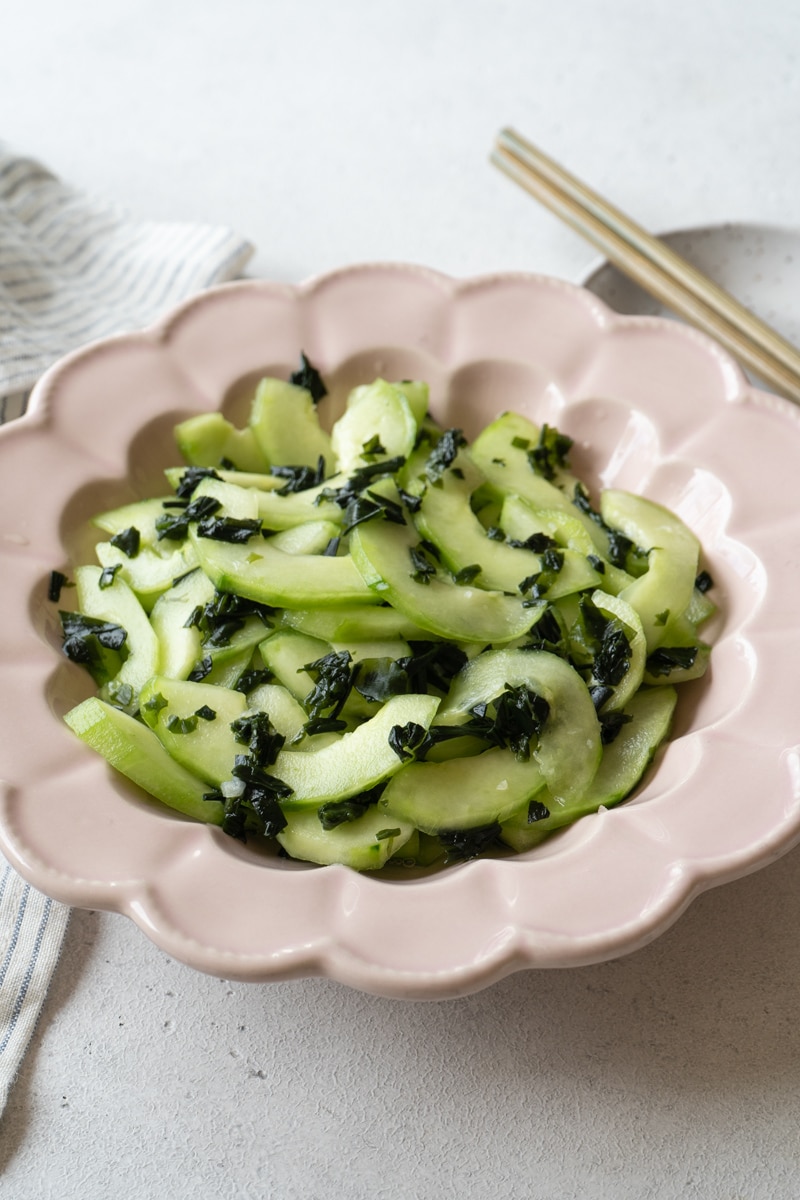 Finished cucumber and seaweed salad in a scalloped pink bowl, garnished with chopped seaweed, with chopsticks resting on a small plate in the background.