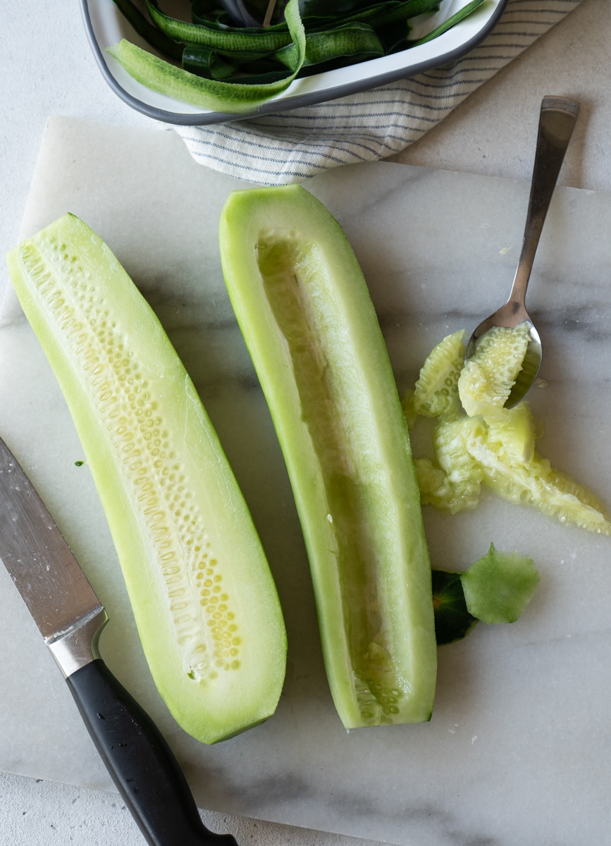 Two cucumber halves with seeds scooped out on a marble board, using a knife and spoon, with more cucumber slices in a bowl in the background."