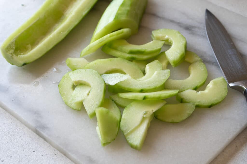 Seeded cucumber halves sliced into thin, crescent-shaped pieces on a marble cutting board next to a knife."