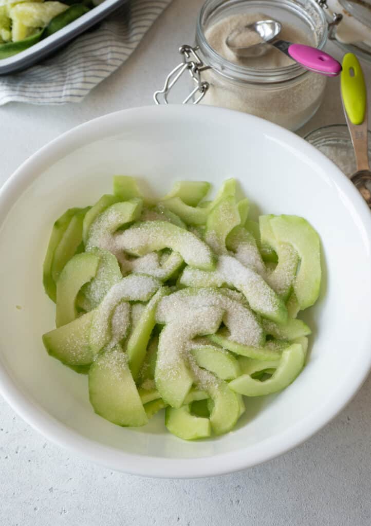 Cucumber slices topped with sugar in a white bowl, with a jar of sugar and measuring spoons in the background.